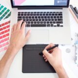 Designer arbeitet mit Laptop und Grafiktablett zwischen Farbmustern und Stiften