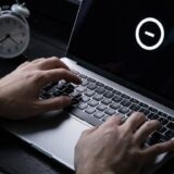 Hände tippen auf einem Laptop mit Ladeanzeige neben einem Wecker